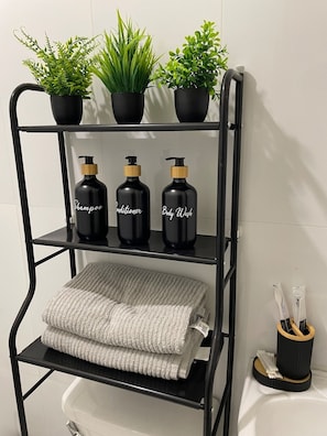 A black metal shelf with three black bottles labeled 'Shampoo,' 'Conditioner,' and 'Body Wash,' and a folded gray towel on the lower shelf. - Cozy Stay at Winland Tower Residences (Quezon City)