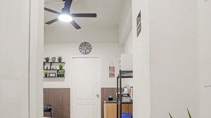 Un pasillo con ventilador de techo, una puerta, una silla azul, una planta en maceta y una zona de cocina con estantes y un refrigerador.