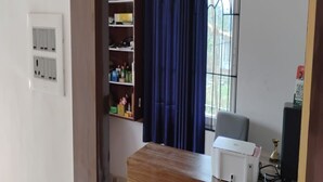A room with a wooden desk, a chair, a printer, a shelf with items, a window with curtains, and electrical outlets. - Village Inn Residency (Kharagpur)