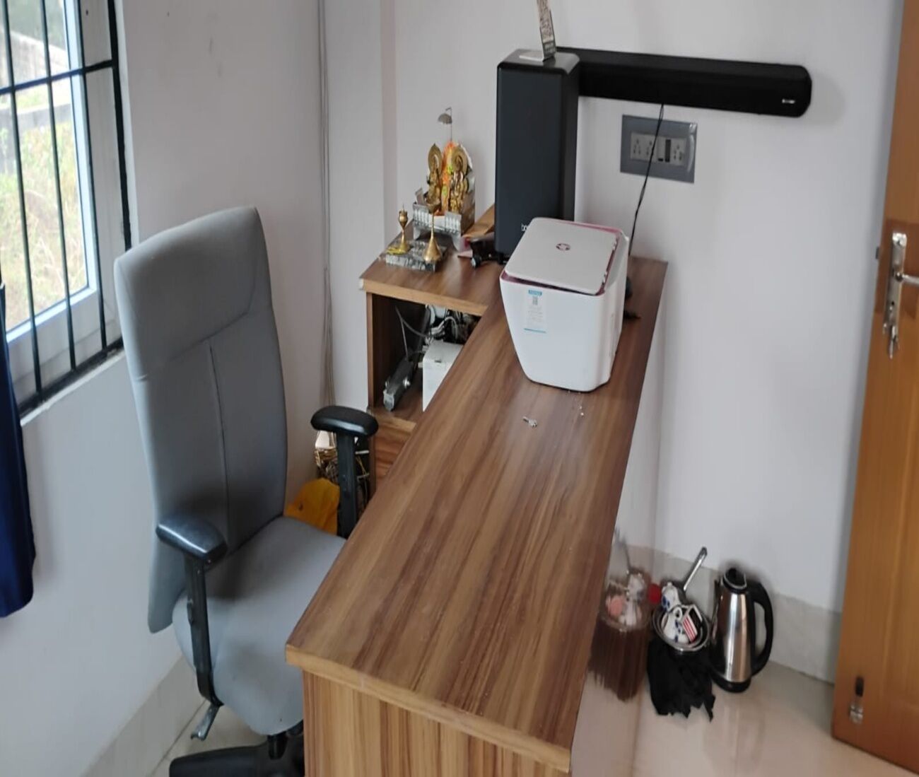 A room with a wooden desk, a chair, a small shelf with decorative items, a white box, a kettle, and a window with bars.