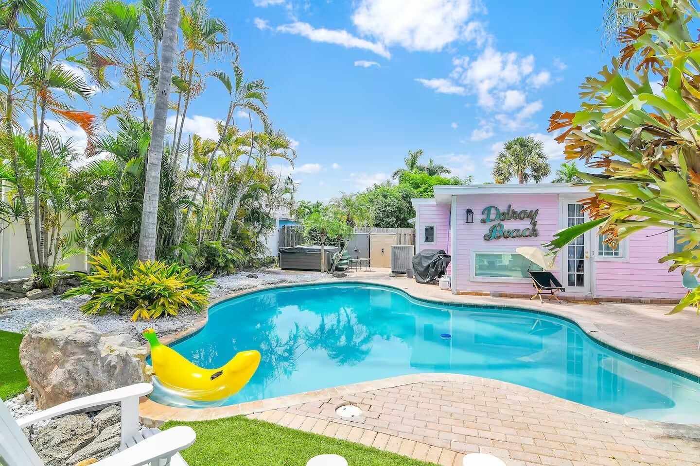 Una piscina con un flotador amarillo, rodeada de palmeras y un edificio rosa con la inscripción 'Delray Beach'.