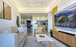 A modern living room with a sofa, a coffee table, a TV, and a kitchen in the background. - City-view 1BR near Junction mall,Gym,pool access (Nairobi)