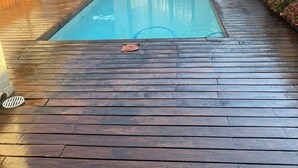 Una piscina con cubierta de madera, una casa con techo de tejas rojas y un pequeño jardín.