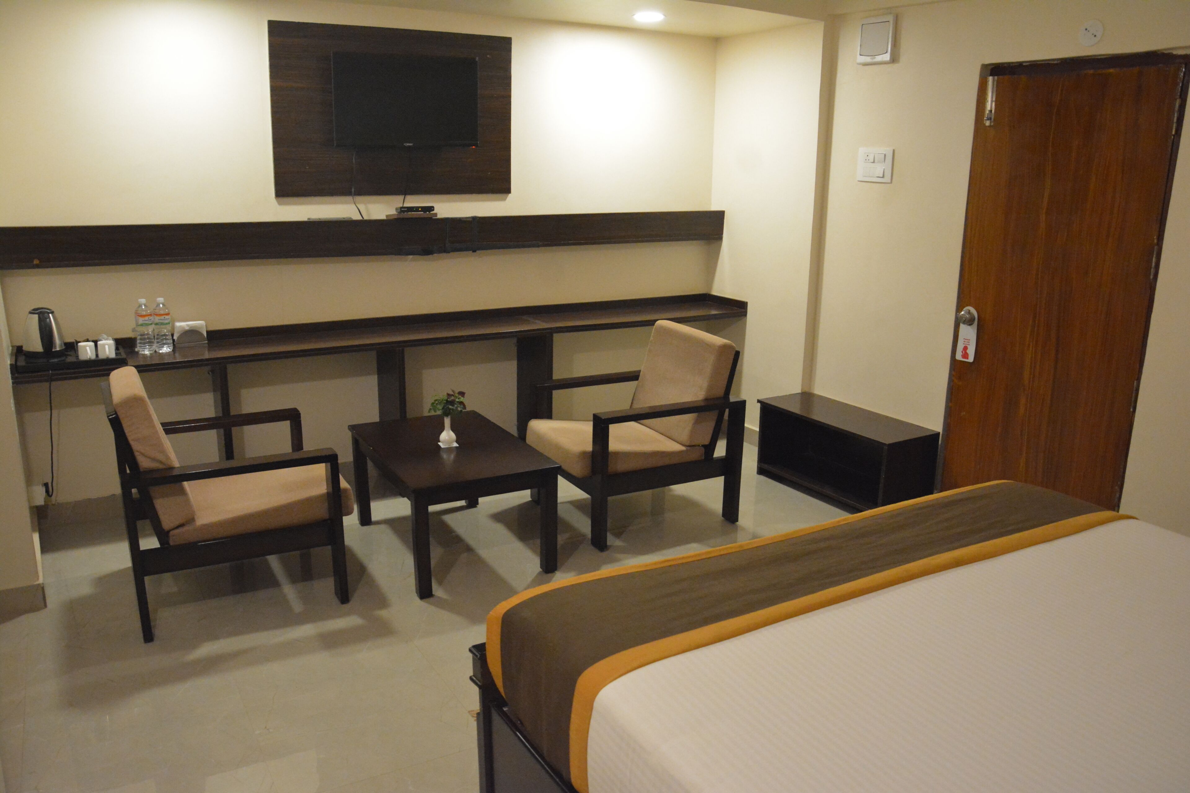Une chambre d’hôtel avec un lit, deux fauteuils, une petite table, un téléviseur fixé au mur et une porte en bois.