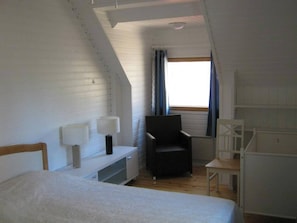 Une petite pièce blanche, avec un toit en pente, comprenant un lit, un bureau, une chaise et une fenêtre avec des rideaux.