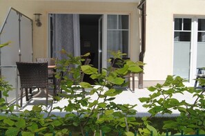 Ein Sitzbereich im Freien mit Korbstühlen und einem Tisch, eine Glastür und ein kleiner Garten mit Grünpflanzen.