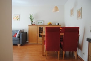 Ein Essbereich mit Holztisch und roten Stühlen, einer Holzkommode, einem grauen Sofa mit Kissen und einer hängenden Pendelleuchte.