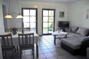 Ein Wohnzimmer mit einer Couch, einem Esstisch mit Stühlen, einem Fernseher und einer Tür, die auf einen Balkon führt.