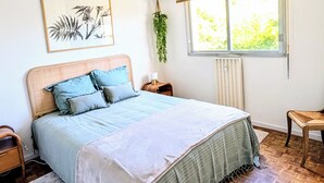 Ein Schlafzimmer mit einem Bett, einem Deckenventilator, einem Fenster mit Jalousien, einem Heizkörper, einer Pflanze und einem Holzboden.