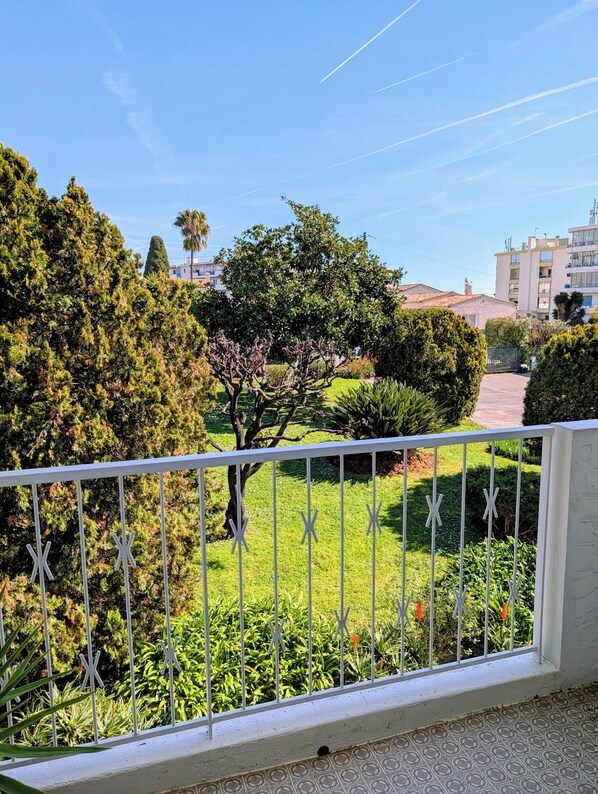 Un balcon avec une balustrade blanche, un jardin bien entretenu avec différents arbustes et un arbre, et un ciel bleu dégagé.