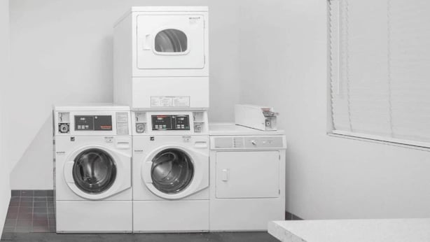 A laundry room with stacked washers and dryers, a small table with a book, and a window with blinds. - Junior Room with Queen Bed|Nsmk (Barstow CA)