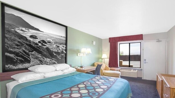 Hotellihuone, jossa on sänky, työpöytä, tuoli, lamppu, ikkuna verhoilla ja suuri kehystetty taulu seinällä.