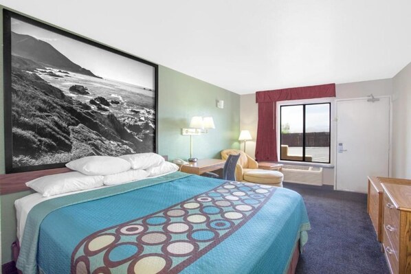 A hotel room with a bed, a desk, a chair, a lamp, a window with curtains, and a large framed picture on the wall.