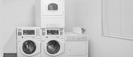 Une buanderie avec des lave-linge et des sèche-linge superposés, une petite table avec un livre et une fenêtre avec des stores.