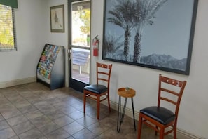Una habitación con dos sillas de madera, una mesa redonda pequeña, un cuadro enmarco con un paisaje de árboles y un tablón de anuncios.