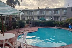 A swimming pool with a poolside area, including a table and chairs, surrounded by a fence and a building in the background.