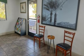 Una habitación con dos sillas de madera, una pequeña mesa redonda, un cartel en la pared y una puerta que da al exterior.
