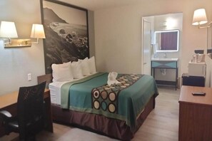 A hotel room with a bed, a desk, a chair, a bathroom with a mirror, and a wall-mounted lamp.