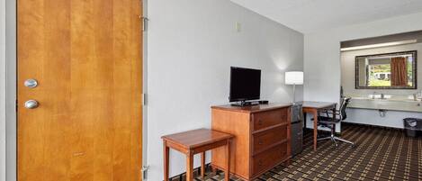 Une chambre d’hôtel avec une porte en bois, un téléviseur posé sur une commode, un bureau avec une chaise, un miroir et un tapis à motifs.