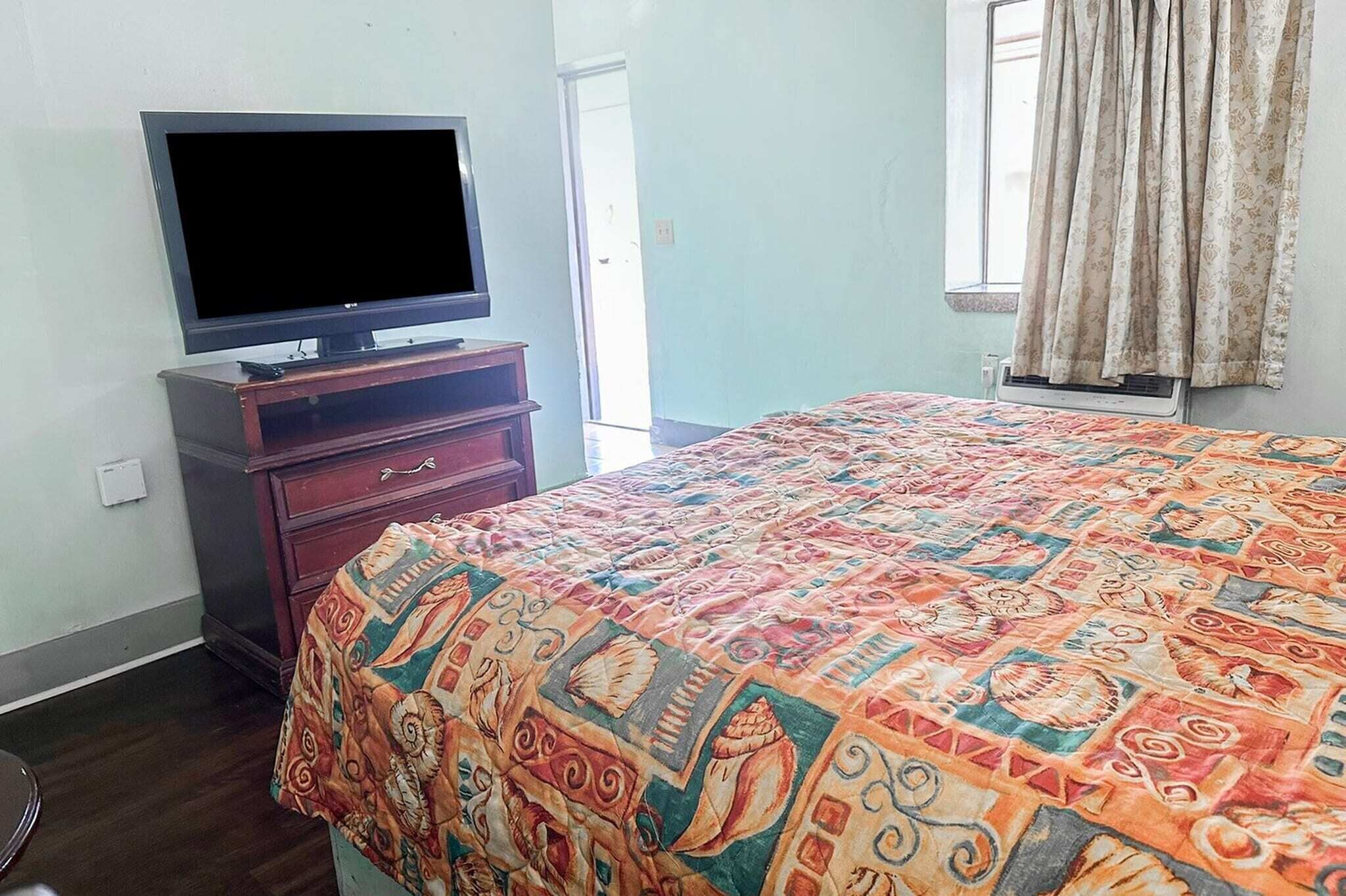 A bedroom with a bed, a TV on a wooden stand, a window with curtains, and a door.