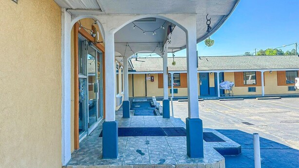 Un espacio exterior techado con suelo de baldosas, un acceso por puerta de cristal y un edificio con fachada amarilla.