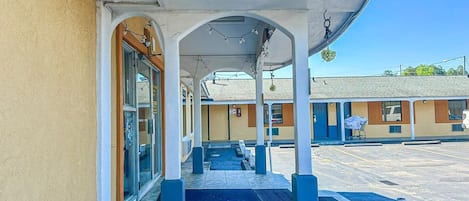 A covered outdoor area with a tiled floor, a glass door entrance, and a building with a yellow facade.