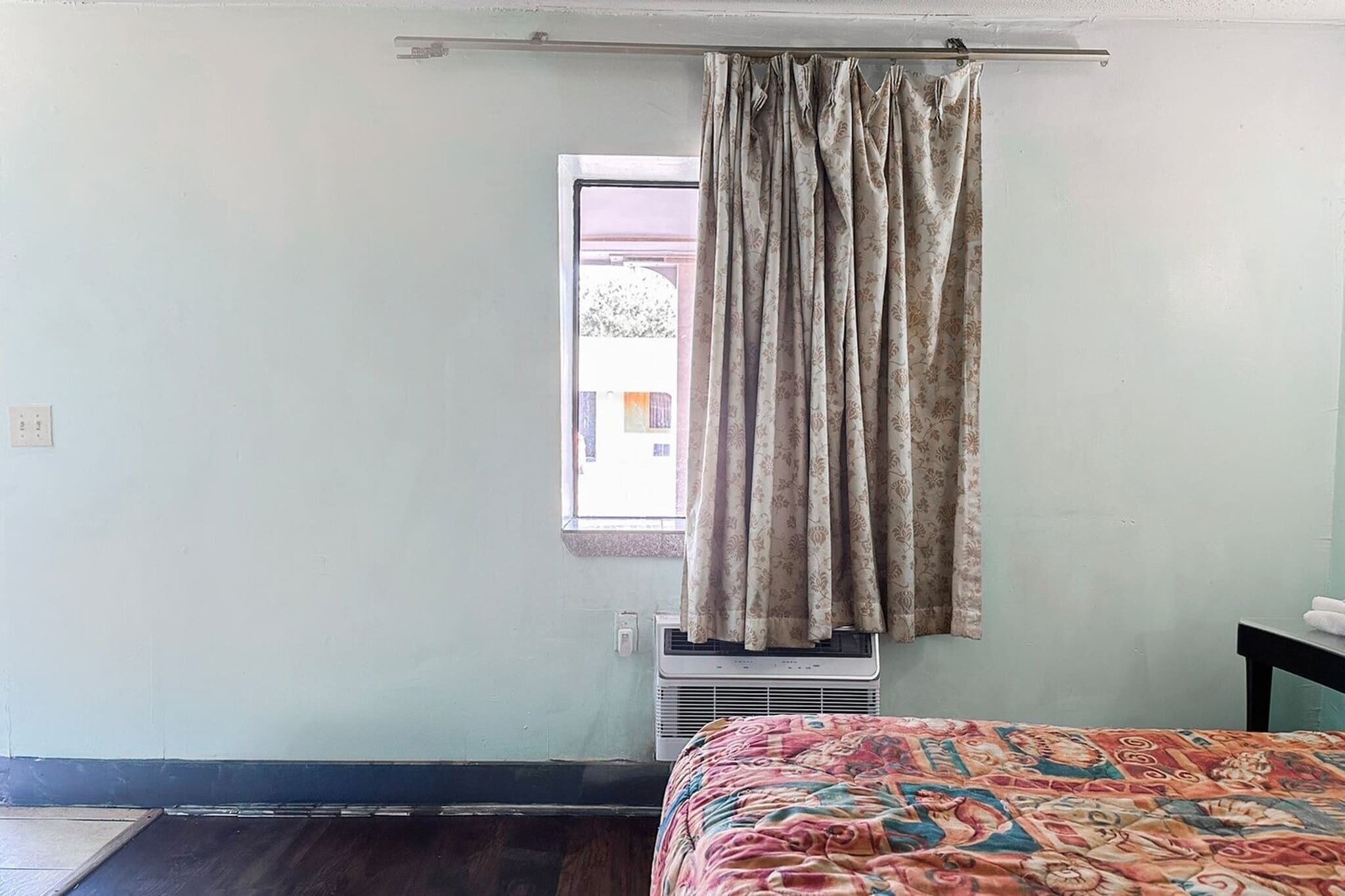 Una habitación con cama, ventana con cortinas, aire acondicionado y vistas a edificios.