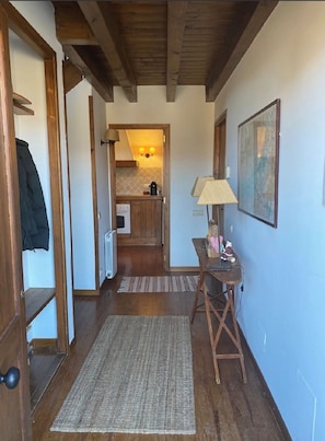 Een gang die leidt naar een keuken met houten balken, een radiator, een tafel met een lamp en een lijst met een afbeelding aan de muur.