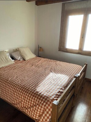 Een eenpersoonsbed met een geruit sprei, een bedlampje en een raam met gordijnen.