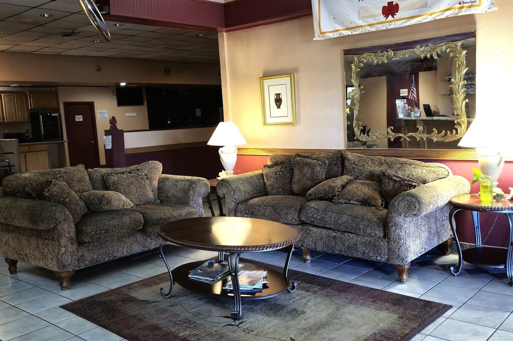 A hotel lobby with two patterned sofas, a round table, a lamp, a mirror, and a kitchen area in the background.