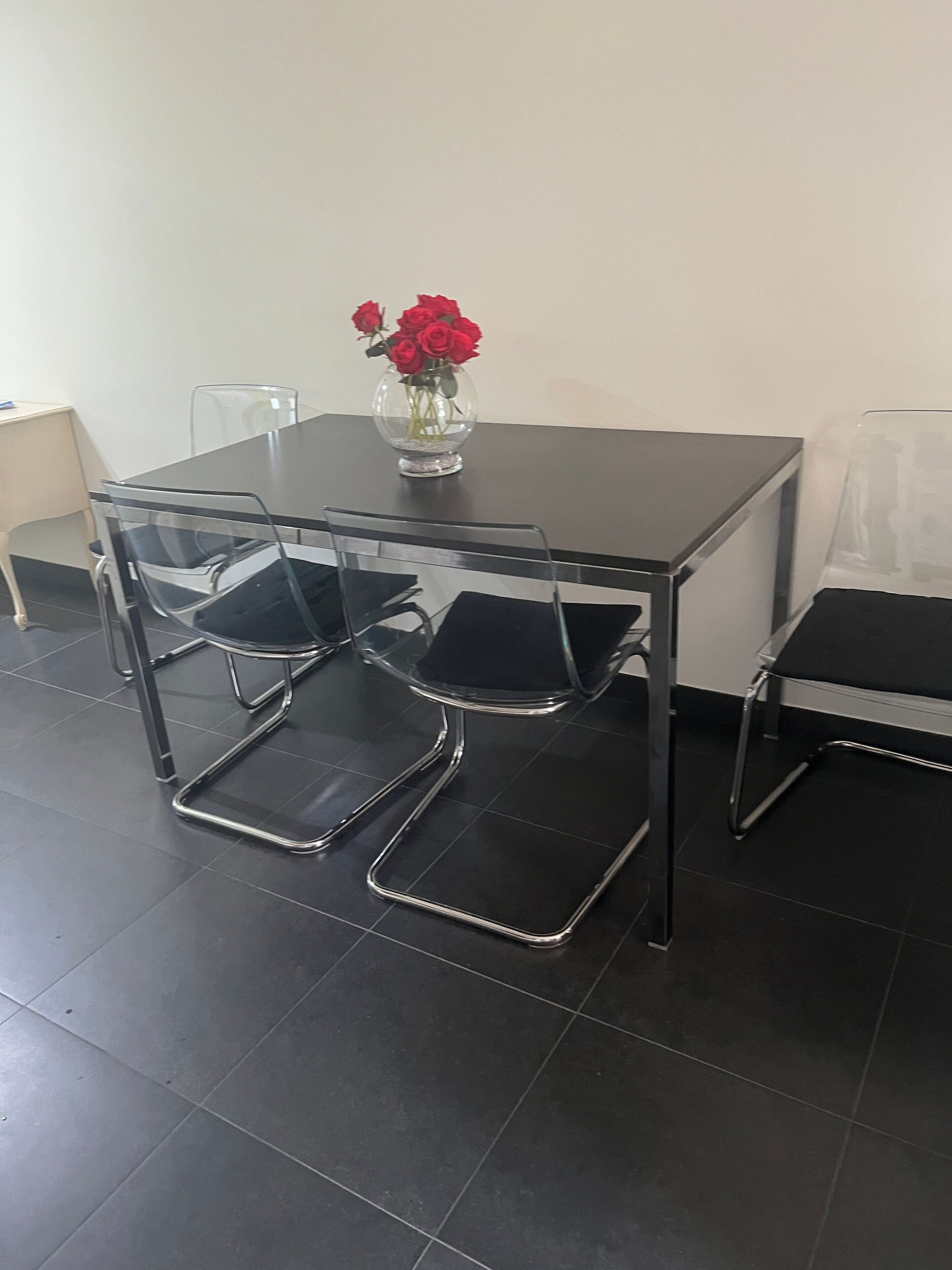 A conference table with four chairs, a vase with red flowers, and a clear plastic chair.