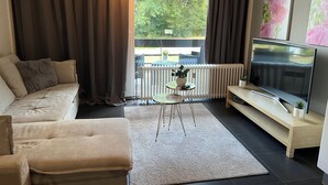 A modern living room with a sofa, a coffee table, a TV, and a view of the outdoors. - Modern Apartment Near Aachen & Dreiländereck (Kelmis)