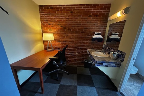 A small, compact room with a wooden desk, a chair, a lamp, a sink, and a mirror.