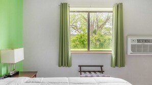 A hotel room with a bed, a nightstand, a lamp, a window with curtains, and an air conditioning unit. - Full Bed|Non-Smoking (Lansing MI)