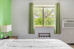 A hotel room with a bed, a nightstand, a lamp, a window with curtains, and an air conditioning unit.
