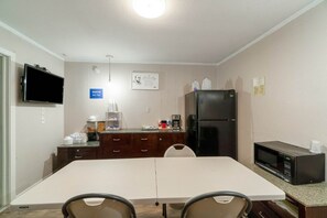 A small kitchenette with a microwave, refrigerator, and coffee maker. There is a dining table with chairs and a TV mounted on the wall.