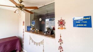 Zona de recepción de un hotel con ventilador de techo, mostrador de recepción, cartel que indica la disponibilidad de desayuno y elementos decorativos en las paredes.