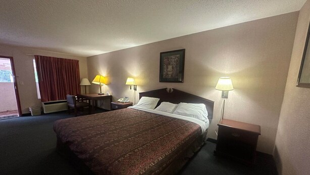 Ein Hotelzimmer mit einem großen Bett, zwei Nachttischlampen, einem Schreibtisch mit Stuhl, einem Fenster mit Vorhängen und einem Wandbild.