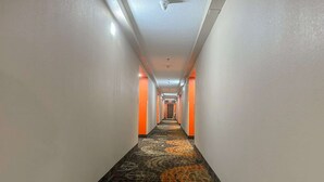 Corredor longo e estreito de um hotel, com carpete estampado, paredes brancas e detalhes em laranja.
