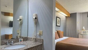 Hotelkamer met een wastafel met granieten blad, twee haardrogerbevestigingen aan de muur, een bed met een bruine quilt en een klein nachtkastje met een magnetron.