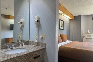 Habitación de hotel con lavamanos de encimera de granito, dos secadores de pelo montados en la pared, cama con colcha marrón y una mesita de noche con microondas.