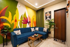 A living room with a blue sofa, a wooden coffee table, a colorful wall mural, a door, and a potted plant.