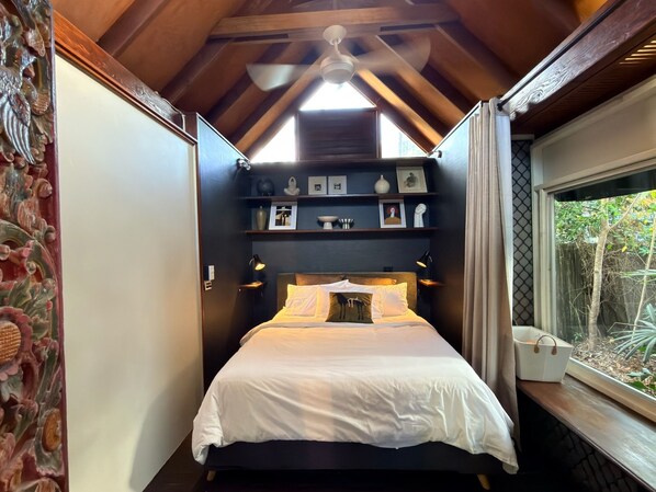 A bedroom with a bed, a ceiling fan, a window with a view of trees, and shelves with various items.