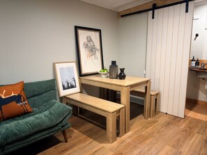 A room with a green sofa, a wooden table with a bench, a framed picture, a vase, and a shelf with toiletries.