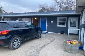 A black SUV parked in front of a building with a blue door, a wooden barrel with flowers, and a small white structure to the right.