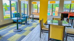 Un hall de hotel con mesas y sillas redondas, ventanas amplias y un pilar amarillo.