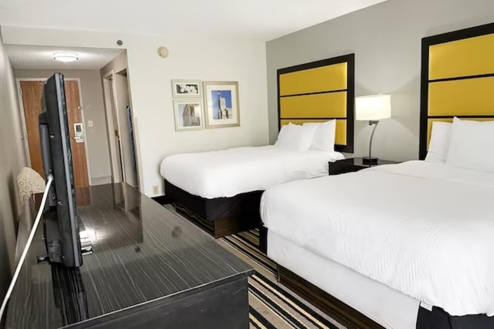 A hotel room with two beds, a flat-screen TV, a bedside table with a lamp, and a framed picture on the wall.