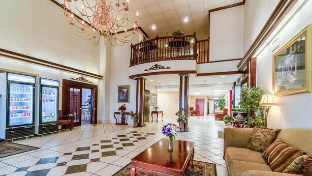 A hotel lobby with a checkered floor, a wooden coffee table with a vase, a sofa, a chandelier, and a balcony. - Suite - King Bed Non - Smoking (Jonesboro, AR)