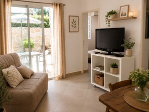 Ein Wohnzimmer mit einer Couch, einem Fernseher auf einem weißen Schrank, einem Esstisch mit Stühlen und Blick auf eine Terrasse.