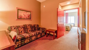 A room with a patterned sofa, a small table, a painting on the wall, and a door leading to a bedroom with beds. - 2 Queen Beds Suite Non-Smoking (Jonesboro, AR)